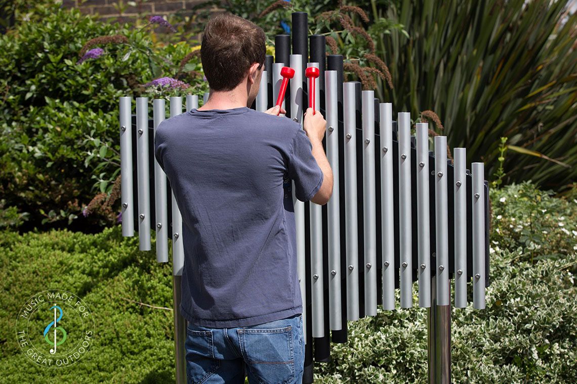 young man playing large outdoor musical instrument with upright tubular notes mounted on black resonators