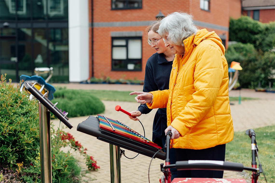 a senior lady wearing a yellow coat standing and playing a rainbow colored outdoor xylophone