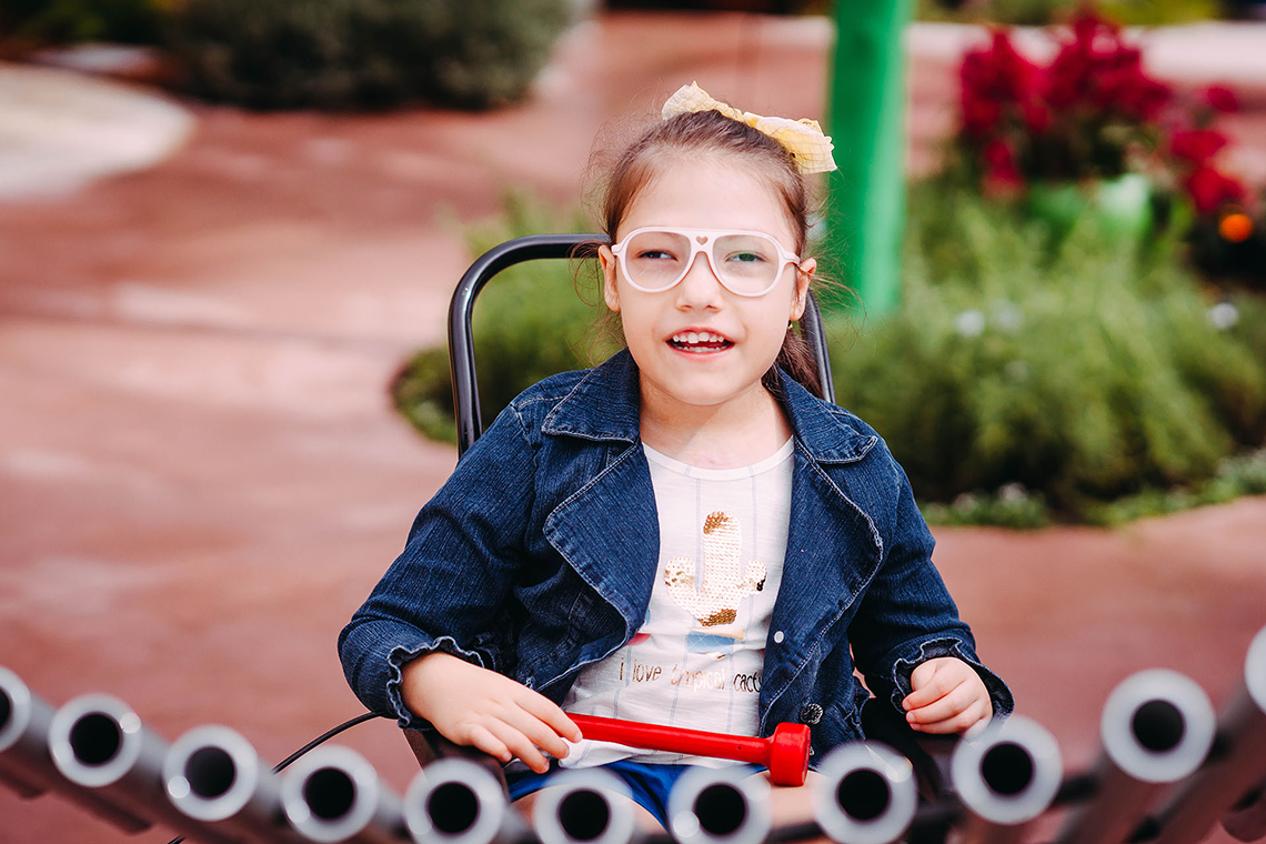 a young girl with special needs and in a wheelchair smiling and playing a large metal outdoor musical instrument in a playground