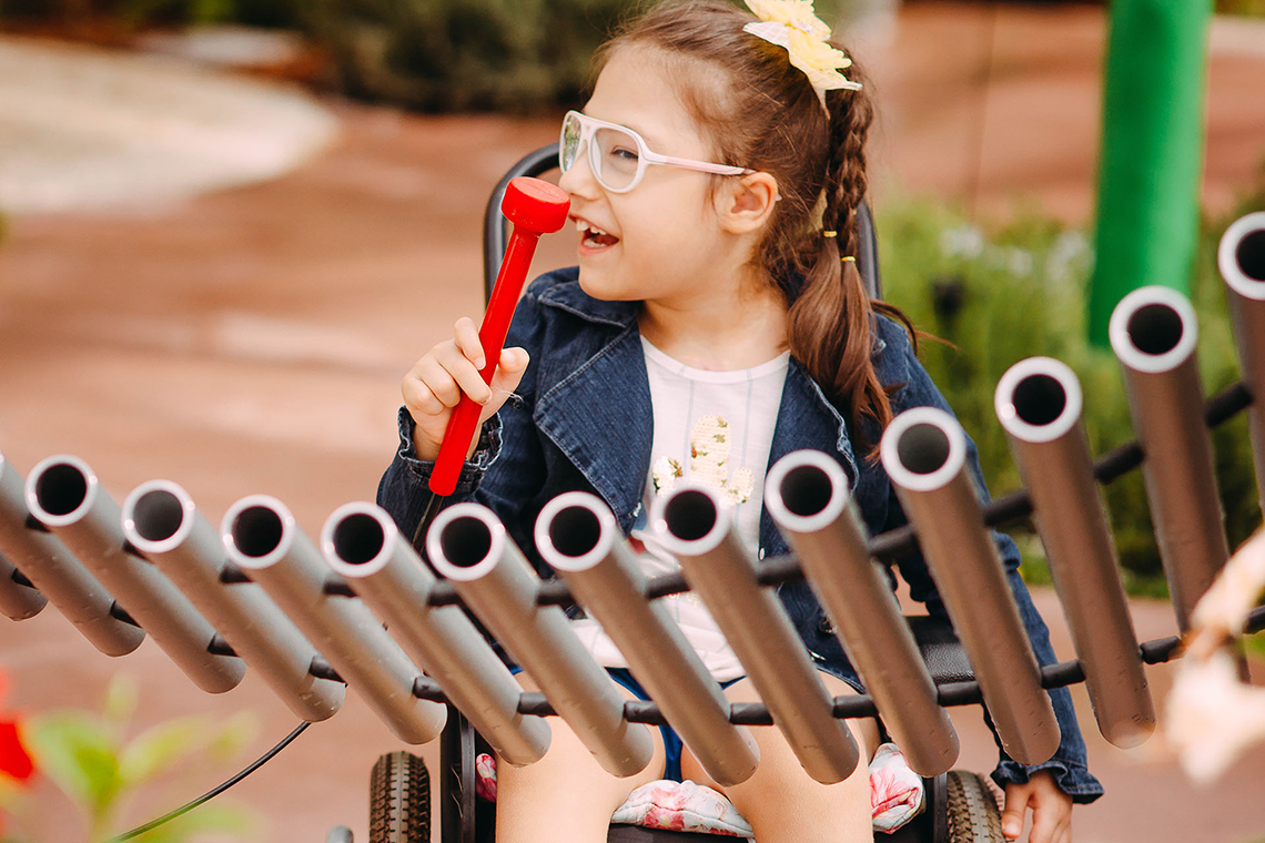 a young girl with special needs and in a wheelchair smiling and playing a large metal outdoor musical instrument in a playground