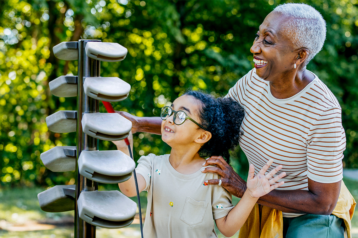 a silver haired older lady of color playing an outdoor musical instrument with her grandadaughter