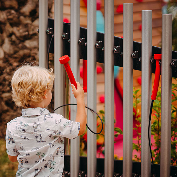 small blonde boy facing away playing a set of large outdoor musical chimes in a music park or playground