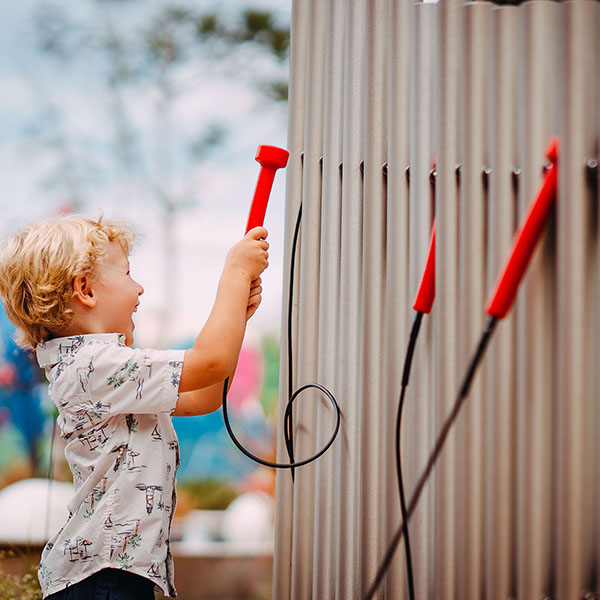 small blonde boy facing away playing a set of large outdoor musical chimes in a music park or playground