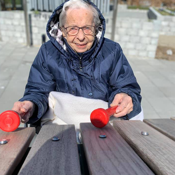 Front on image of an older lady in a wheelchair playing the wooden notes of a large outdoor musical instrument