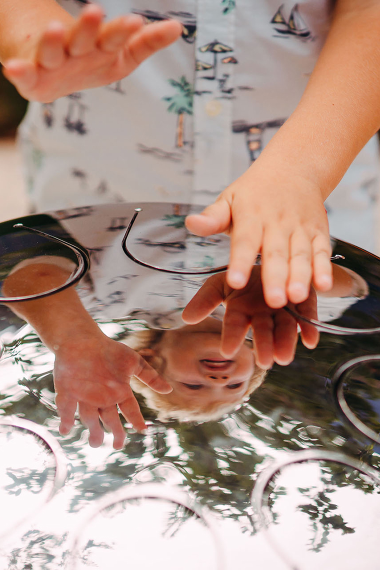 a stainless steel tongue drum in a musical playground or park reflecting the hands of its young player