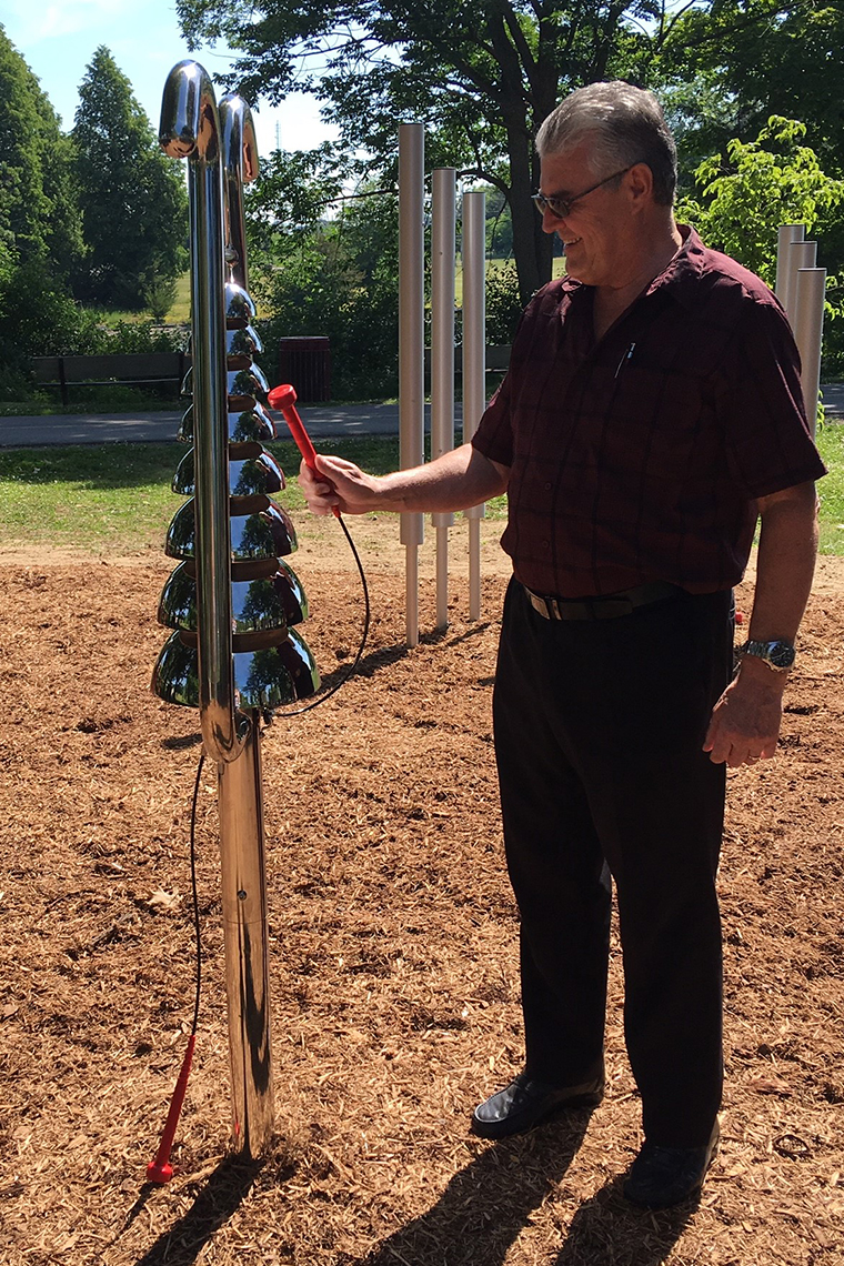 a senior man playing an large outdoor musical instrument shaped like a bell tree in a rotary music garden