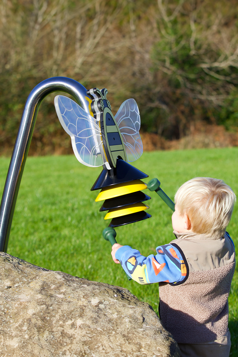 an outdoor musical instrument shaped like a bumblebee on a stainless steel post being played by a young blond boy