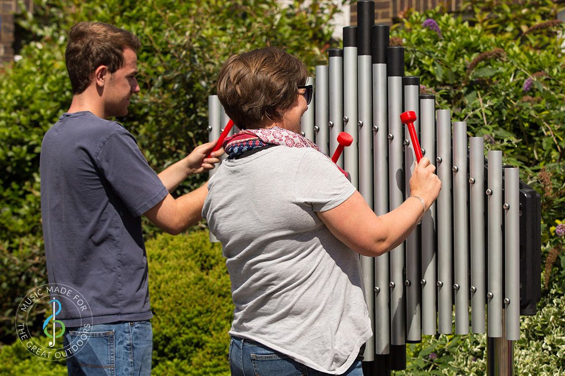 two people playing large outdoor musical instrument with upright tubular notes mounted on black resonators