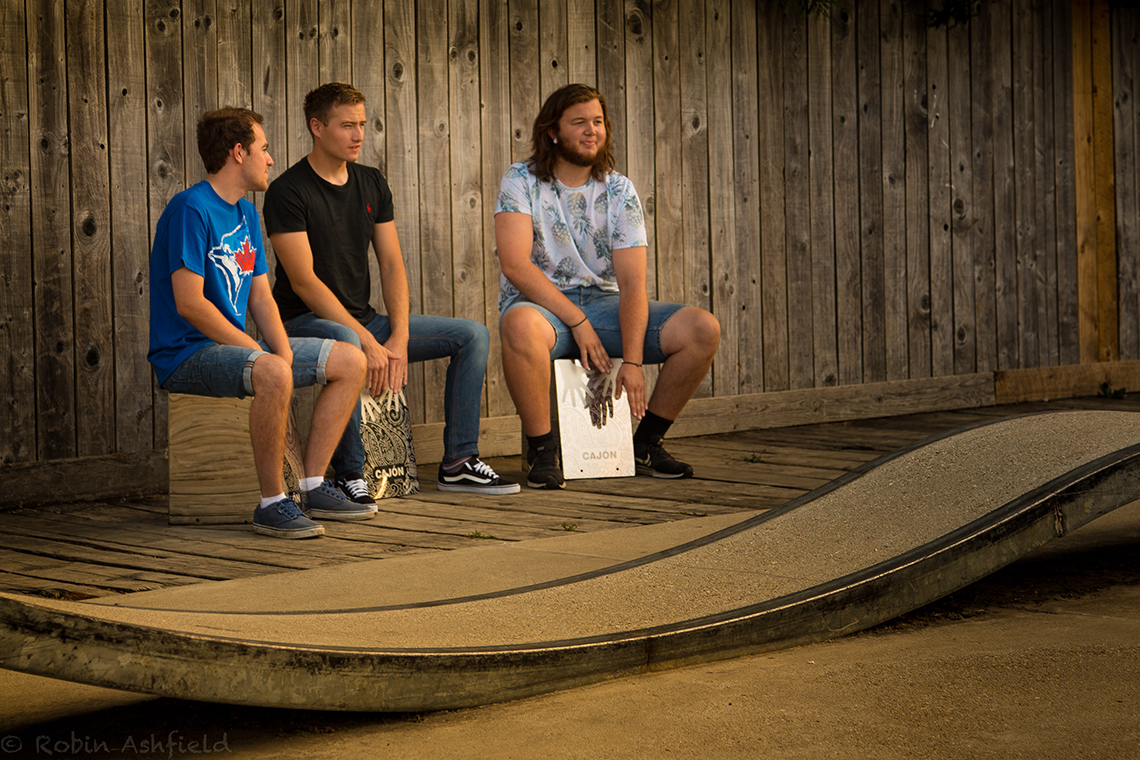 three young men sitting on stainless steel cajon percussion drums in an outdoor shelter