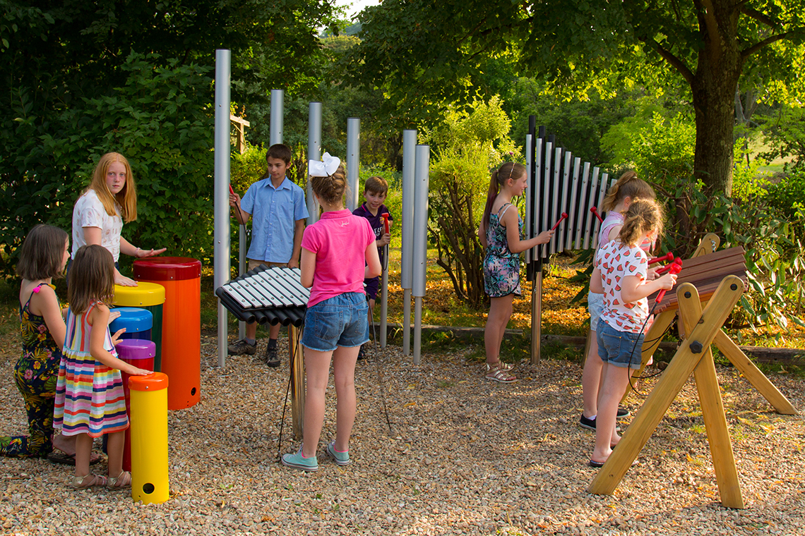 large group of children playing on an ensemble of outdoor musical instruments in a park