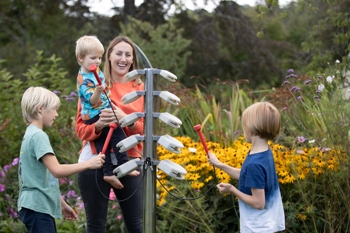 a mother holding a baby and with two young boys playing on an outdoor musical instrument shaped like a tree with branches made of temple blocks to be hit with red mallets